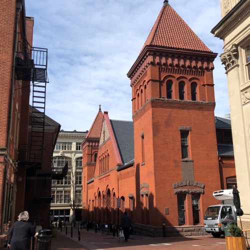 Historic red brick building with a pointed roof, surrounded by modern architecture and a clear blue sky.