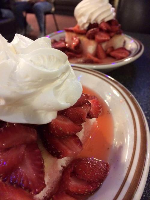 A close-up of a dessert topped with whipped cream and fresh strawberries on a plate, with another dessert in the background.