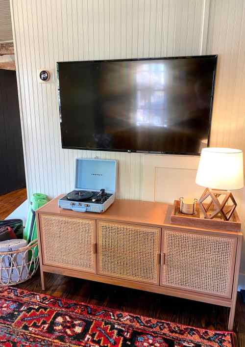 A living room scene featuring a TV mounted on the wall, a record player on a wooden cabinet, and a lamp.