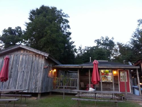 A rustic wooden building with red umbrellas outside, surrounded by trees and a grassy area. Warm lights glow from the windows.