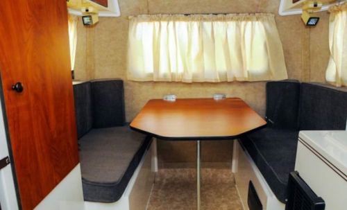 Cozy interior of a camper with a wooden table and cushioned seating, illuminated by natural light through curtains.