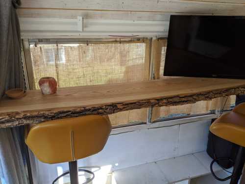 A wooden countertop with a natural edge, two bar stools, and a TV in a cozy, sunlit space with sheer curtains.