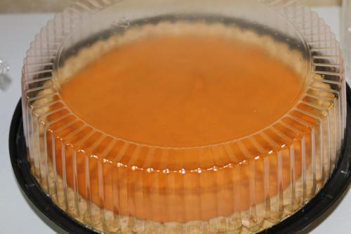 A round dessert with a smooth orange topping, covered by a clear plastic dome, resting on a black base.