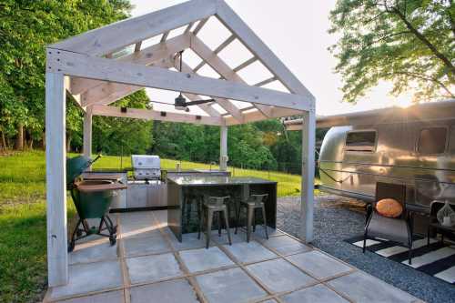 Outdoor kitchen with a grill, countertop, and seating under a wooden structure, beside a vintage Airstream trailer.