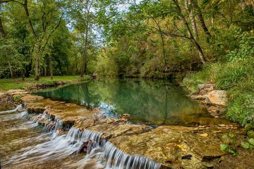 A serene river scene with gentle waterfalls, surrounded by lush green trees and a calm, reflective pool.