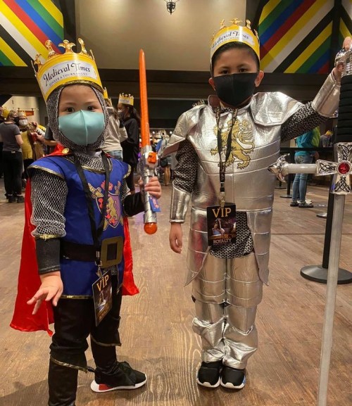 Two children dressed as knights, one in a blue and red costume with a cape and the other in silver armor, both wearing crowns.