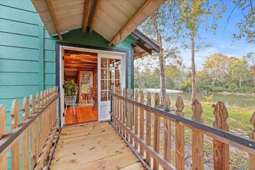 A wooden walkway leads to a charming green house with a view of a river and trees in the background.