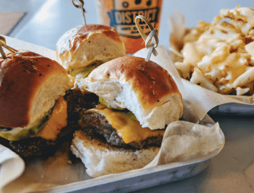 Three mini cheeseburgers with pickles on a tray, alongside a serving of cheesy fries and a drink in the background.