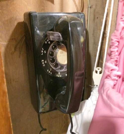 A vintage black rotary phone mounted on a wooden wall, with a pink curtain in the background.