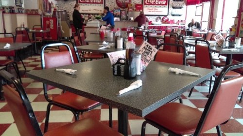 A diner interior with empty tables, red and white checkered floor, and a counter in the background.