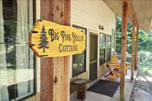 Wooden sign reading "Big Pine Hollers Cottage" on a porch with a rocking chair and greenery in the background.