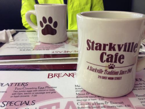 Two coffee mugs on a table with a menu, one mug features a paw print, and the other reads "Starkville Cafe."