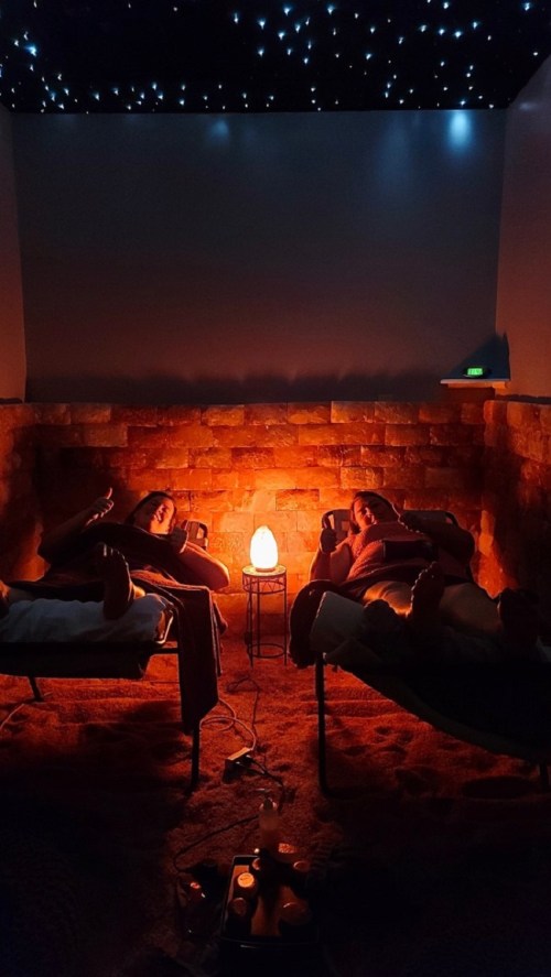Two people relax on lounge chairs in a dimly lit room with a salt wall and a glowing salt lamp.