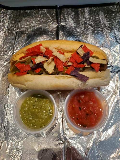 A hot dog topped with colorful tortilla strips, served with two small cups of green salsa and tomato salsa.