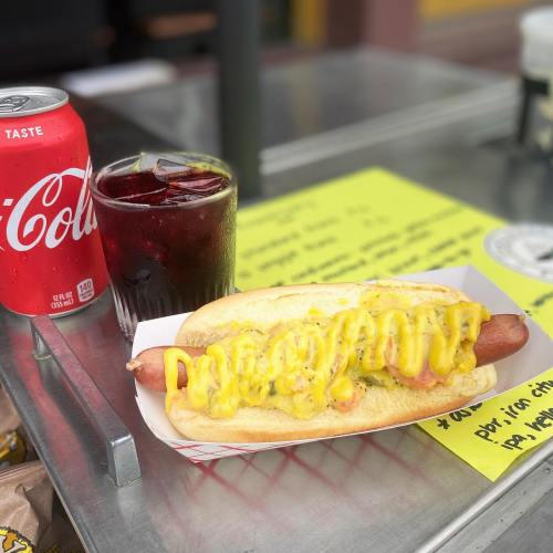 A hot dog topped with mustard and a drink beside it, set on a table with a colorful menu in the background.