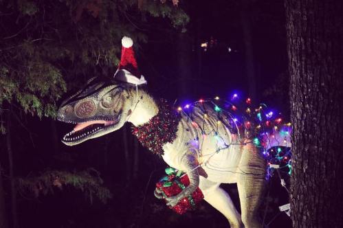 A dinosaur statue wearing a Santa hat, adorned with colorful lights and a festive garland, stands in a wooded area.