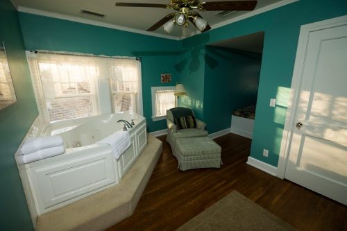 A cozy bathroom featuring a large tub, a green accent wall, a comfortable chair, and natural light from windows.