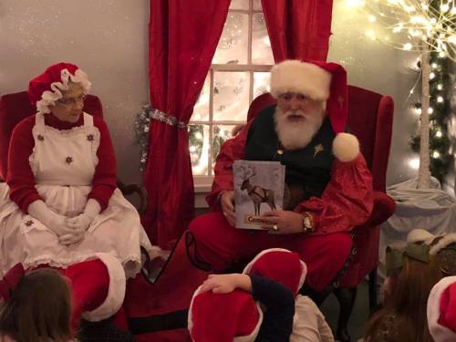 Santa Claus reads a book to children while sitting next to Mrs. Claus in a festive, decorated room.