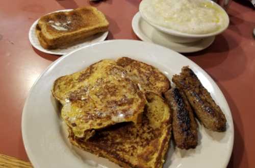 A plate with French toast, sausage links, and a bowl of grits on a red table. A slice of toast is on a small plate.