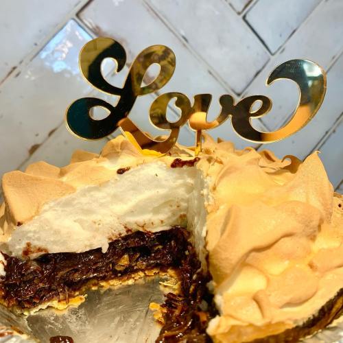 A chocolate cake with a creamy topping, featuring a gold "Love" cake topper and a slice cut out.