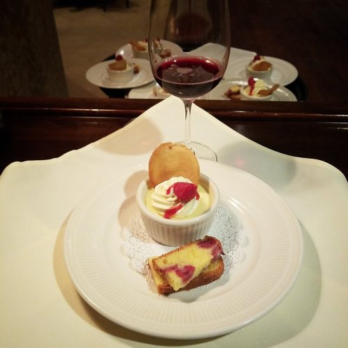 A dessert plate with creamy dessert topped with a raspberry, a slice of cake, and a glass of red wine in the background.