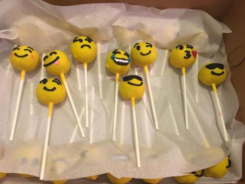 A tray of yellow cake pops decorated with various emoji faces, including happy, sad, and playful expressions.