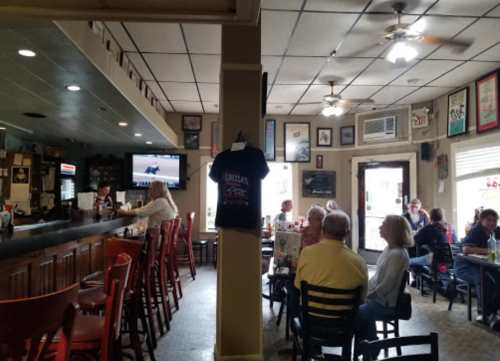 A cozy bar scene with patrons seated at tables and the bar, a TV showing a race, and various framed pictures on the walls.