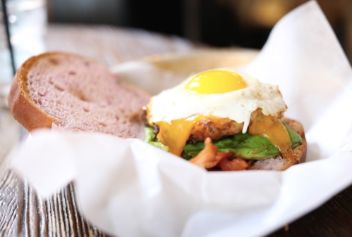 A close-up of a sandwich with a fried egg, cheese, spinach, and tomato on brown bread, served on a wooden table.