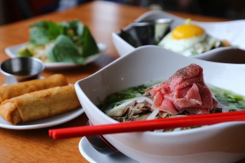 A bowl of pho topped with sliced meat, green herbs, and spices, with spring rolls and other dishes in the background.