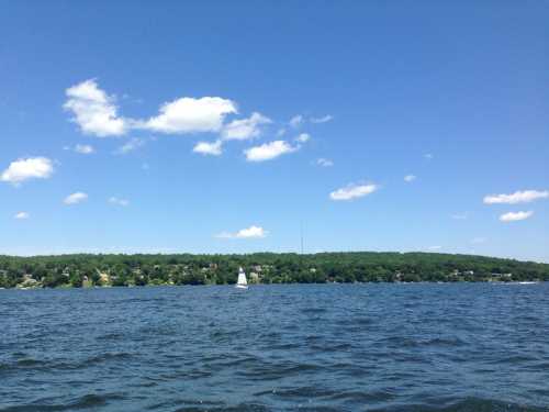 A serene lake scene with a sailboat, green hills, and a clear blue sky dotted with fluffy white clouds.