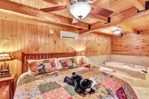 Cozy wooden bedroom with a quilted bed, bear plush toy, and a jacuzzi tub, featuring warm lighting and rustic decor.