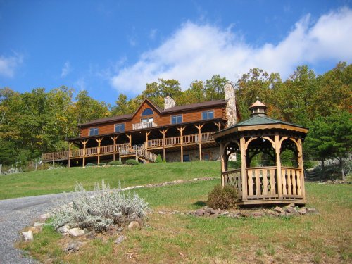 A large wooden house on a hillside, with a gazebo in the foreground and trees in the background under a blue sky.