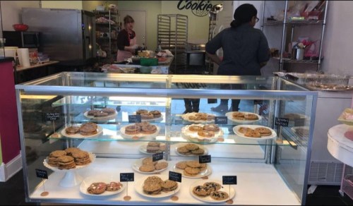 A display case filled with various cookies, with a baker working in the background of a cozy bakery.