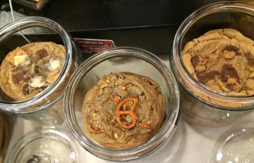 Three jars filled with large, colorful cookies featuring various toppings like chocolate chunks and pretzels.