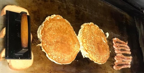 Two golden pancakes and strips of bacon cooking on a griddle, with a spatula nearby.