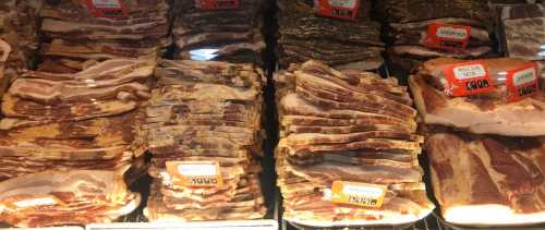 A display of various packaged bacon strips in a grocery store, labeled with price tags.