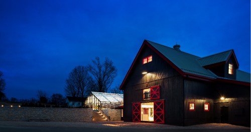 A dark barn with red accents stands beside a glass greenhouse, illuminated against a twilight sky.
