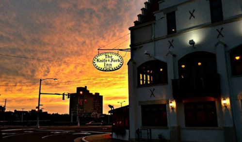 The Knife-Fork Inn sign illuminated against a vibrant sunset sky, with a cityscape in the background.