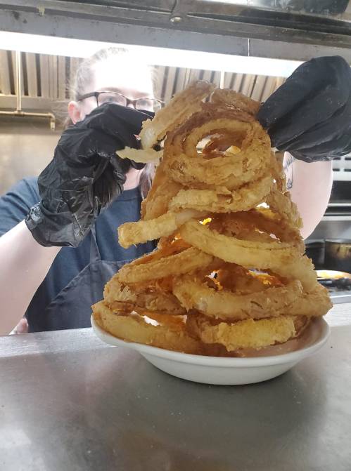 A person wearing black gloves holds a tall stack of crispy onion rings above a bowl in a kitchen setting.