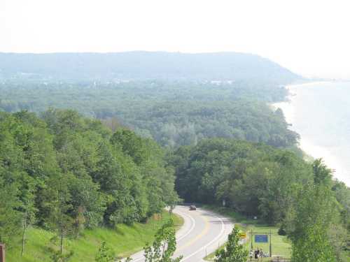 A scenic view of a winding road surrounded by lush greenery, leading to a coastline and distant hills under a hazy sky.