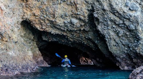 A kayaker paddles through a rocky cave entrance, surrounded by water and textured stone walls.