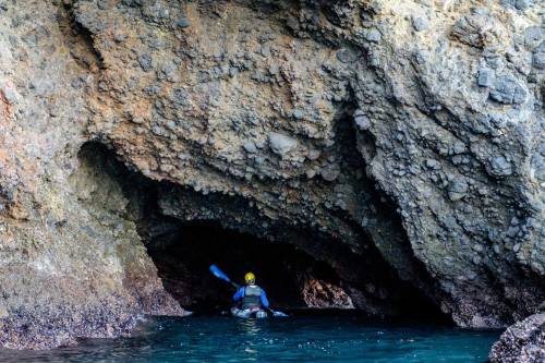 A person in a kayak paddles through a rocky cave entrance, surrounded by water and textured stone walls.