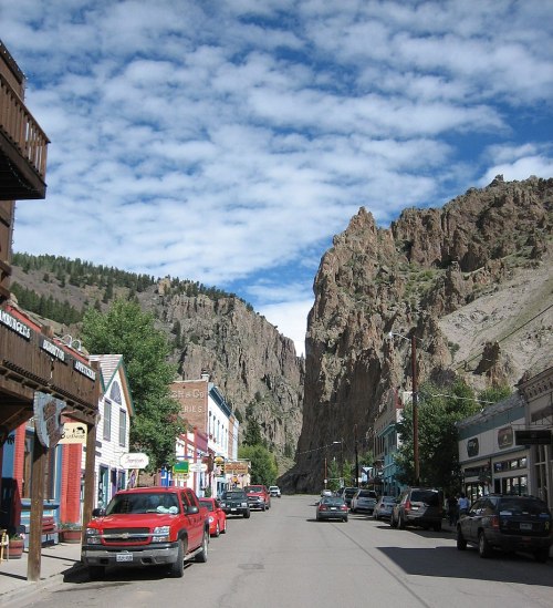 A narrow street lined with colorful buildings and parked cars, flanked by rocky cliffs under a partly cloudy sky.