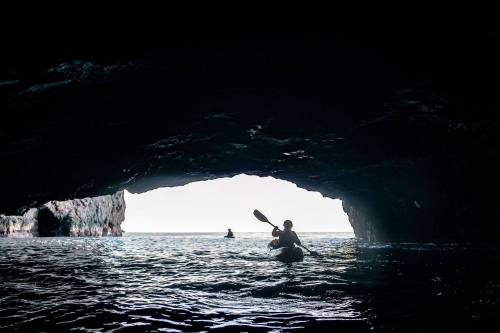 A person kayaking through a dark cave, with another kayaker visible in the distance on the water.