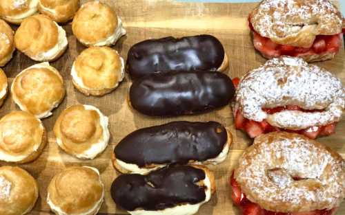 A wooden board displaying an assortment of pastries: cream-filled puffs, chocolate eclairs, and strawberry croissants.