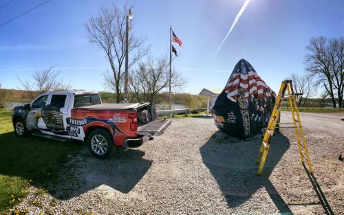 A pickup truck with patriotic graphics parked near a large, flag-themed structure and a ladder on a gravel area.