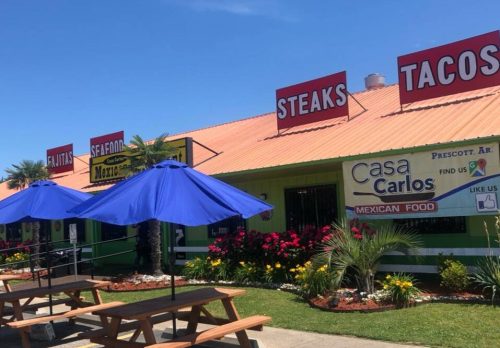 Colorful restaurant exterior with signs for tacos, steaks, and Mexican food, featuring outdoor seating and palm trees.