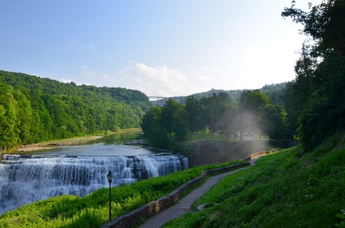 A serene landscape featuring a waterfall, lush greenery, and a pathway along the river under a clear blue sky.