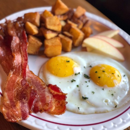 A plate with two sunny-side-up eggs, crispy bacon, golden potato cubes, and apple slices.