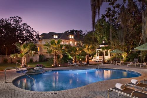 A serene pool area with palm trees, lounge chairs, and a gazebo, set against a twilight sky and a beautiful house.
